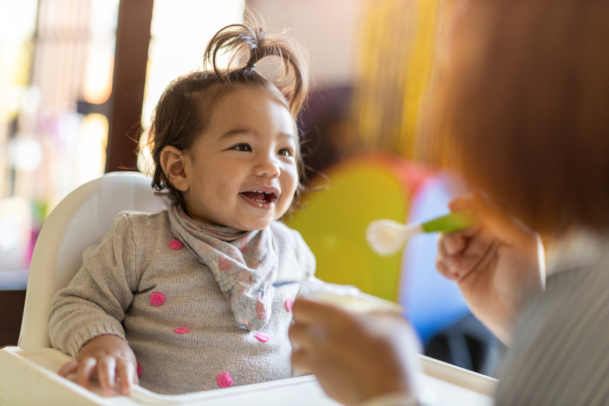 Picture of a baby in a high chair smiling with a woman holding a jar of baby food and a spoon